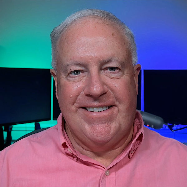 Headshot of Chuck Joiner sitting in front of his podcast workstation. Three monitors in the background with blue and green background lit from behind.